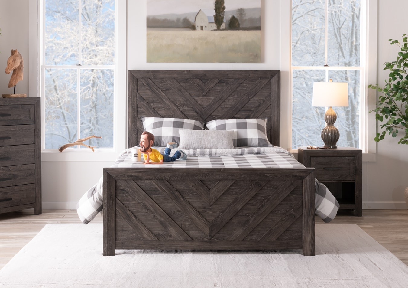 Cozy dark gray wooden bedroom set with chevron patterned bed, 5 drawer chest, and nightstand with drawer and a double gourd lamp, anchored on a rustic white carpet with plaid pillows and blanket.