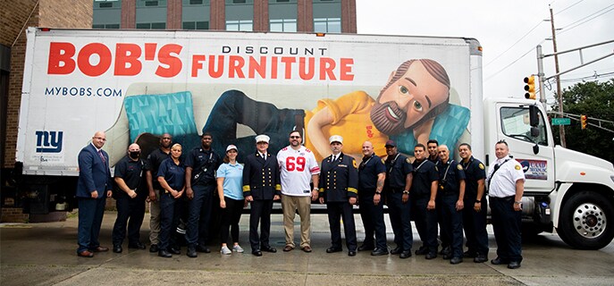 Several Jersey City Fire Department firefighters standing in front of Bob's furniture truck with New York Giants Legend Rich Seubert
